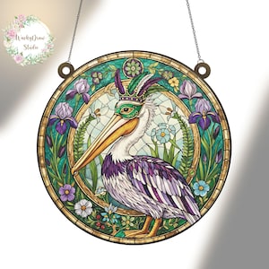Op de afbeelding: Ronde glas-in-lood raam met een pelikaan met masker en kroon, omringd door bloemen en bladeren. De pelikaan heeft paarse en witte veren, een gele snavel en een turquoise en gouden achtergrond. Inclusief ophangketting.