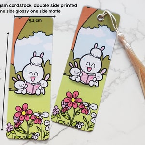 Könnte beinhalten: Zwei doppelseitige Lesezeichen mit einem Comic-Kaninchen, das ein Buch mit anderen Kaninchen liest. Die Lesezeichen sind 5,2 cm breit und 15,2 cm hoch. Die Lesezeichen haben einen grünen Hintergrund mit rosa Blumen und einem weißen Quasten.