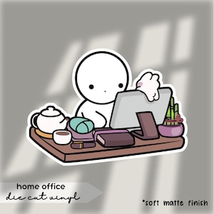 Op de afbeelding: Een gestanste vinylsticker met een minimalistische illustratie van een persoon aan een thuiskantoor. Het bureau bevat een computer, een theeset, boeken en een kleine plant. De tekst "home office" en "die cut vinyl" zijn opgenomen.