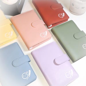 May include: A collection of colorful, leather-like journals in various shades including red, pink, green, blue, yellow, and purple. Each journal features a flap closure and a white graphic of a smiling face with a pen. The journals are arranged on a white surface.
