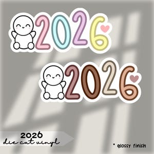 Op de afbeelding: Twee gestanste vinylstickers met het jaar 2026. Eén sticker heeft pastelroze, blauwe en gele cijfers met een hartje en een lachend cartoonfiguur. De andere heeft bruine en beige cijfers met een hartje en een lachend cartoonfiguur. Tekst "2026 die cut vinyl".