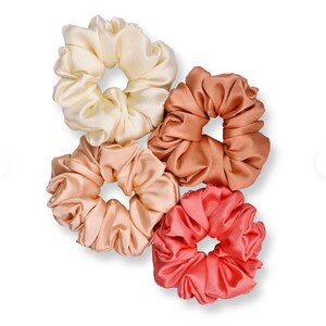 Può includere: Quattro scrunchies di raso in tonalità pesca, corallo e crema. Gli scrunchies sono disposti in un gruppo su uno sfondo bianco.