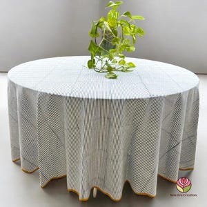May include: A circular tablecloth featuring a white backdrop with navy blue vertical stripes. The edge of the cloth has a yellow trim. A glass vase with green plants is placed on the tablecloth.