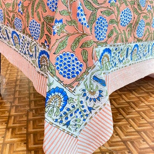 Puede incluir: Un mantel de mesa de color melocotón con un patrón floral en tonos de azul, verde y blanco. El mantel tiene un borde blanco con un patrón floral azul y verde.