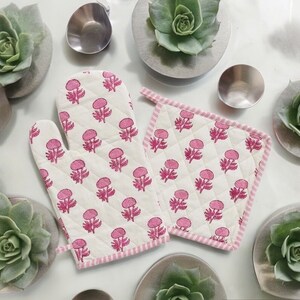 Könnte beinhalten: Ein Set aus zwei rosa und weißen Baumwoll-Topflappen mit Blumendruck. Die Topflappen sind gesteppt und haben einen rosa Saum.