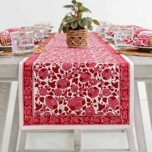 May include: A red and white floral patterned table runner. The runner features a repeating design of stylized flowers and leaves. A small potted plant sits in the centre of the table. The table is set with glasses and napkins.