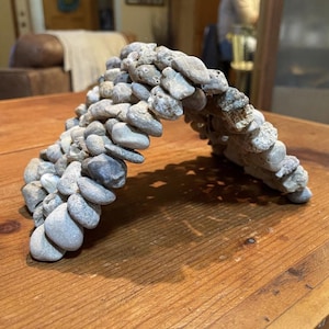 May include: A handcrafted stone archway made of various sized, light gray river rocks. The arch is constructed in a curved shape and sits on a wooden surface. The natural stone arrangement creates a rustic, decorative element.