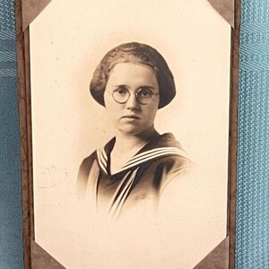 May include: A black and white photograph of a woman wearing round glasses and a sailor-style shirt. She has short, wavy hair and is looking directly at the camera.