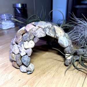 May include: A handcrafted stone archway, constructed from various sized, multi-colored river rocks. The arch is designed for terrarium or vivarium use, providing a naturalistic habitat element. Air plants are placed around the arch.
