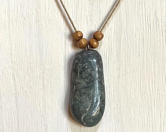 Op de afbeelding: Een ketting met een grijze stenen hanger met een natuurlijke, gladde afwerking. De ketting is gemaakt van een eenvoudig bruin koord en houten kralen.