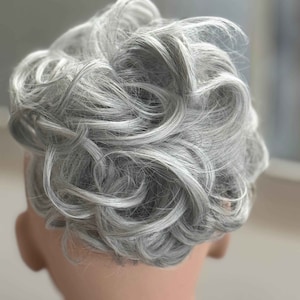 May include: A close-up of a wig with short, wavy, gray hair. The hair is styled in a voluminous, curly fashion, with the curls cascading around the head. The wig is on a mannequin head.