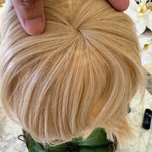 May include: A blonde wig with a circular opening at the crown, showing the base of the wig. The wig is styled in a layered, slightly messy look.