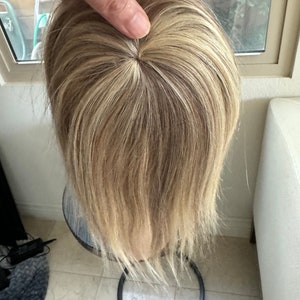 Könnte beinhalten: Ein blonder Topper aus Echthaar mit einer runden Öffnung an der Krone. Der Topper steht auf einem schwarzen Ständer.