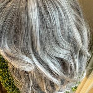 May include: A long, wavy, gray wig with a natural-looking parting. The wig is styled in loose waves and has a light gray color.