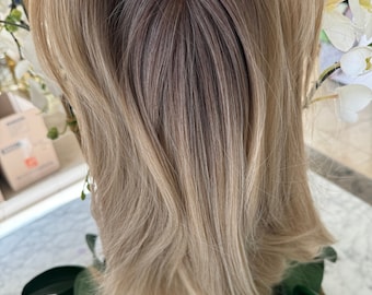 May include: A light brown wig with blonde highlights. The wig is styled in a long, layered cut with a centre parting. The wig is made of human hair and is designed to look natural.