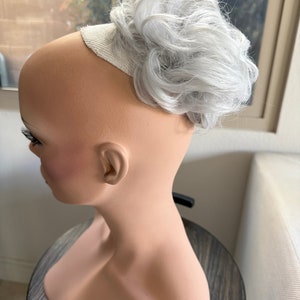 May include: A silver grey hairpiece in a messy bun style. The hairpiece is attached to a white elastic band.
