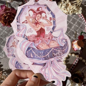 May include: A die-cut sticker featuring a jester character with a red and white hat sitting inside an open purple coffin. The character wears a red corset and tutu, surrounded by pink and blue ribbons, pearls, and small floating teeth.