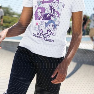 Puede incluir: Camiseta blanca con un gráfico estilo manga. El texto dice "HUNTR" y "KPOP DEMON HUNTERS". El gráfico presenta tres paneles con ilustraciones de estilo anime en morado, azul y rosa. La persona lleva pantalones negros.