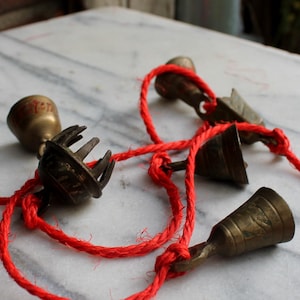 Può includere: Una collezione di piccole campane antiche in ottone infilate su un cordino rosso brillante. Le campane variano per forma e dimensione, con dettagli intricati. Il cordino rosso è annodato in più punti. Le campane sono disposte su una superficie di marmo bianco e grigio.