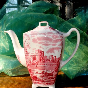 Op de afbeelding: Een witte keramische theepot met een rood kasteelontwerp. De theepot heeft een gebogen tuit, een handvat en een deksel met een klein handvat. Het ontwerp toont een kasteel, bomen en wolken. De theepot staat op een houten oppervlak.