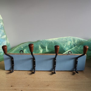 Può includere: Un attaccapanni rustico da parete con quattro ganci. Il supporto presenta una base in legno azzurro chiaro invecchiato con ganci in metallo scuro e pomelli in legno marrone. I ganci sono distanziati e pronti all'uso.