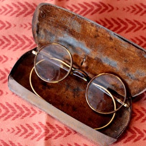 Może przedstawiać: Okulary w okrągłej oprawie w stylu vintage ze złotymi oprawkami spoczywają w otwartym, zużytym drewnianym etui. Etui jest w kolorze ciemnobrązowym, a okulary mają przezroczyste soczewki. Tło to wzorzysta czerwona tkanina.