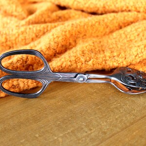 Puede incluir: Pinzas de servir de plata con un diseño decorativo y adornado en el mango. Las pinzas están sobre una superficie de madera.