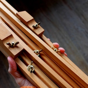 Könnte beinhalten: Ein hölzernes Künstlerstaffelei mit verstellbaren Holzstützen und Messingbeschlägen. Die Staffelei wird von einer Person gehalten, was ihr kompaktes, faltbares Design zeigt. Die natürliche Holzfarbe und die Messingakzente verleihen ihr ein klassisches Aussehen.