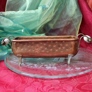 Puede incluir: Un recipiente rectangular de cobre martillado con acabado metálico, que cuenta con cuatro pequeñas patas de metal plateado y dos asas curvas en los laterales, cada una acentuada con un pequeño detalle de cerámica blanca en el extremo.
