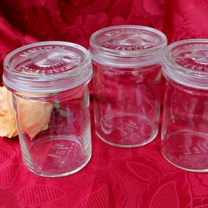 Op de afbeelding: Drie heldere glazen potten met deksels, geplaatst op een rode stof met patroon. De potten hebben een vintage uitstraling, met reliëftekst op de deksels. Een lichtgele roos ligt naast een van de potten.