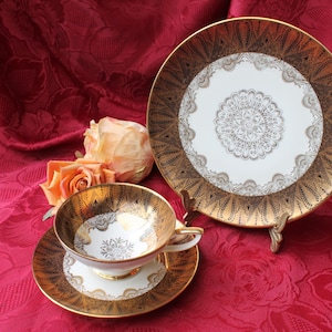 Peut inclure: Un service à thé vintage avec un motif doré et blanc. L'ensemble comprend une tasse à thé, une soucoupe et une assiette décorative. La tasse et la soucoupe sont placées devant l'assiette, qui est posée sur un petit support. Le fond est un tissu floral rouge.
