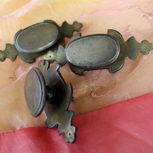 Op de afbeelding: Antieke messing ladetrekjes met ovale middenplaten en decoratieve achterplaten. De hardware heeft een vintage uitstraling, met een aangetaste afwerking. De trekjes zijn geplaatst tegen een roze en crème stoffen achtergrond.