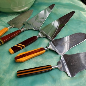 Könnte beinhalten: Vier Tortenheber aus Metall mit dekorativen Griffen in verschiedenen Streifenmustern in Gelb, Orange, Braun und Schwarz. Die Edelstahlklingen haben unterschiedliche Formen, darunter dreieckige und abgerundete Kanten, teilweise mit floralen Gravuren.