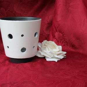 Könnte beinhalten: Ein weißer Keramiktopf mit schwarzem Rand und Boden, der mehrere kreisförmige Aussparungen aufweist. Eine weiße Rose liegt neben dem Topf auf einem roten Stoff mit floralem Muster. Der Topf ist etwa 10 cm hoch.