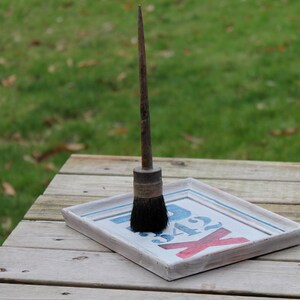 Könnte beinhalten: Ein Vintage-Pinsel mit langem Holzgriff und schwarzen Borsten steht auf einem gerahmten Schild. Das Schild hat einen weißen Hintergrund mit blauen und roten Buchstaben. Die Szene spielt sich auf einem verwitterten Holztisch im Freien ab.