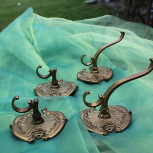 Peut inclure: Quatre patères murales antiques en laiton avec des plaques arrière ornées d'inspiration florale et des crochets incurvés. Les crochets sont disposés sur un fond en tissu turquoise, mettant en valeur leur design vintage.