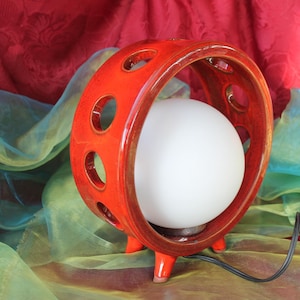 Könnte beinhalten: Eine Lampe im Retro-Stil mit einem leuchtend orangefarbenen Keramikrahmen. Der Rahmen hat kreisförmige Aussparungen und hält eine weiße Kugellampe. Die Lampe hat drei kleine Beine und ein schwarzes Netzkabel.