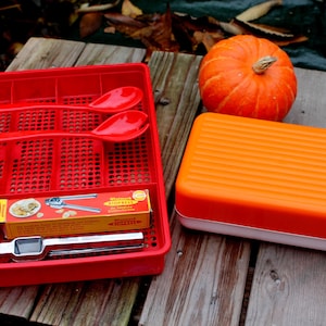 Könnte beinhalten: Ein roter Küchenutensilien-Organizer mit geschlitzten Abschnitten, der rote Servierlöffel, einen Nussknacker und ein Thermometer enthält. Ein oranger Kürbis und ein rechteckiger orange-weißer Behälter liegen auf einer Holzoberfläche.