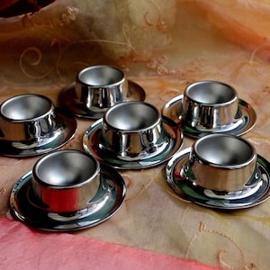 Peut inclure: Six coquetiers argentés brillants avec leurs soucoupes assorties sont disposés sur un tissu pêche à motifs. Les coquetiers ont un design classique et reflètent la lumière. Les soucoupes sont rondes et légèrement concaves.