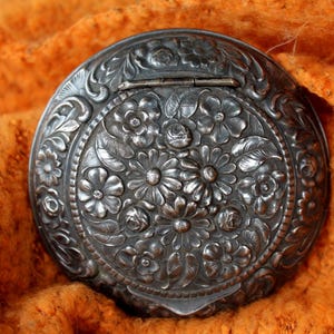 Puede incluir: Un compacto de plata con un intrincado diseño floral. El compacto tiene una tapa con bisagras y un patrón floral.