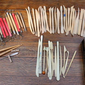 Op de afbeelding: Een verzameling aardewerkgereedschap, waaronder houten modelleerstaafjes, draadlussen met rode en houten handvatten, metalen kogelstylussen en een draadsnijder. Rechts staat een zwarte rechthoekige doos met gouden tekst en vogelillustraties.