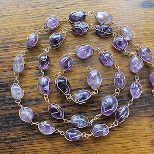 Op de afbeelding: Een lange ketting gemaakt van amethiststenen die zijn gewikkeld in gouden draad. De stenen zijn diep paars van kleur en hebben allemaal verschillende vormen en maten. De ketting is geregen op een gouden ketting.