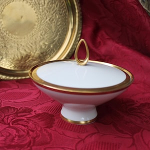 Könnte beinhalten: Weiße Porzellan-Zuckerdose mit goldfarbenem Rand und einem goldenen Knopf auf dem Deckel. Die Schale steht auf einer roten, floralen Tischdecke.