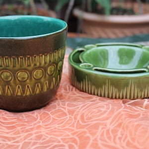Può includere: Un set di vasi e posacenere in ceramica. Il vaso è marrone con interno verde e un motivo ripetuto di cerchi e zig-zag. Il posacenere è verde con esterno a coste e diversi supporti.
