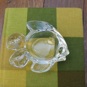 Könnte beinhalten: Eine klare Glasschale in Fischform mit ausgeprägten Flossen, einem Schwanz und einem kreisförmigen Becken in der Mitte. Das Glas ist transparent und glatt und weist geformte Details auf, die die Anatomie des Fisches nachzeichnen, inklusive eines kleinen Auges.