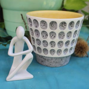 Pode incluir: Uma estatueta de cerâmica branca de uma pessoa em posição de joelhos ao lado de um vaso de cerâmica. O vaso apresenta um exterior creme com um padrão repetitivo de reentrâncias cinzentas texturizadas e uma borda interior amarela.