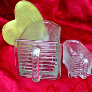 Peut inclure: Deux pichets en verre transparent avec des poignées, un grand et un petit, sont posés sur une surface rouge. Les pichets ont une forme rectangulaire avec des lignes horizontales. Un objet jaune en forme de cœur est en arrière-plan.