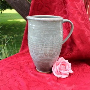 Op de afbeelding: Een lichtgrijze keramische kan met een handvat, die op een rode stof met een bloemenpatroon staat. Een roze roos ligt naast de kan.