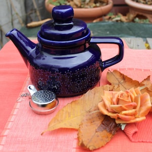 Op de afbeelding: Een donkerblauwe keramische theepot met bijpassende deksel, handvat en tuit. De theepot heeft een textuurpatroon en staat op een rode placemat. Een roestvrijstalen thee-infuser en een oranje roos met bladeren zijn ook aanwezig.