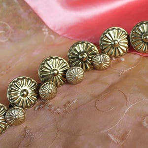 Könnte beinhalten: Eine Sammlung goldfarbener, dekorativer Knöpfe in verschiedenen Größen. Die Knöpfe haben ein Sonnenstrahl-Design und sind auf einem rosa- und pfirsichfarbenen Stoff mit einem dezenten Muster angeordnet. Die Beleuchtung hebt den metallischen Glanz der Knöpfe hervor.